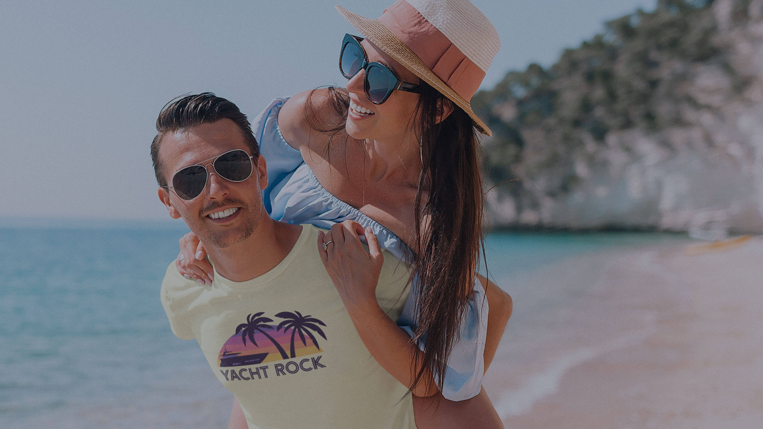 A woman is smiling as she is carried on a man's back at the beach while he is wearing the "A Yacht Rock Sunset" tee shirt by Yacht Rock Clothing.